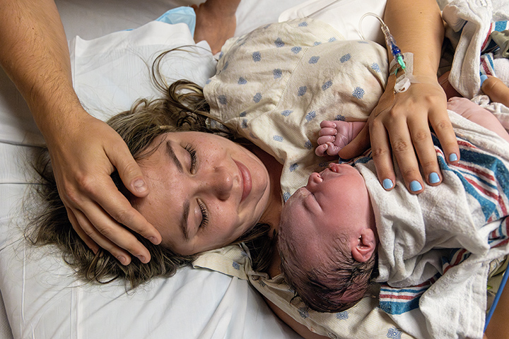 woman holding newborn baby while husband puts his hand on her head in hospital bed setting