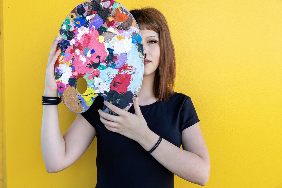 high school student on yellow backdrop holding up painting supplies